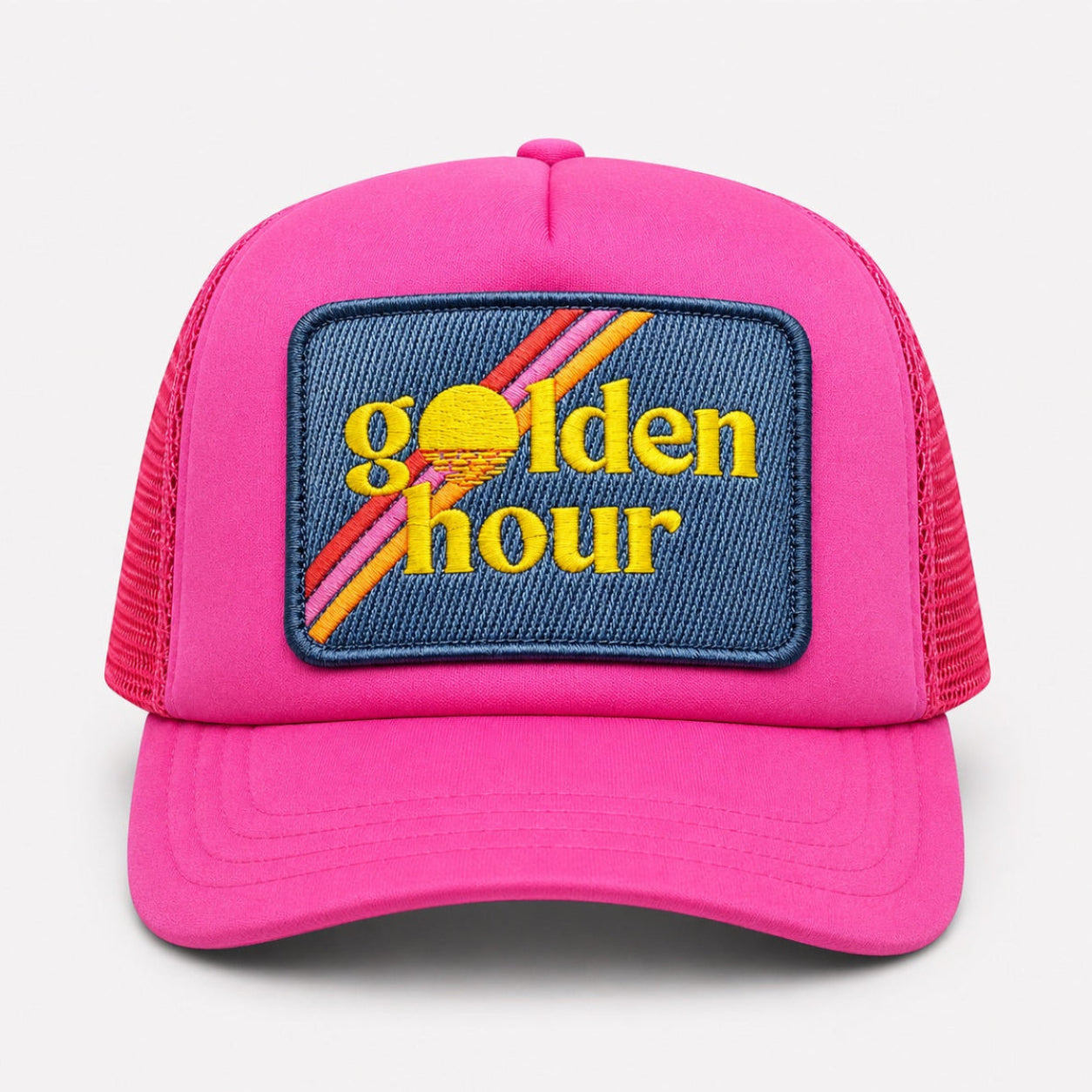 A bright pink trucker hat with a blue embroidered patch on the front that reads 'golden hour' in yellow with diagonal orange and red stripes, shown against a light gray background.